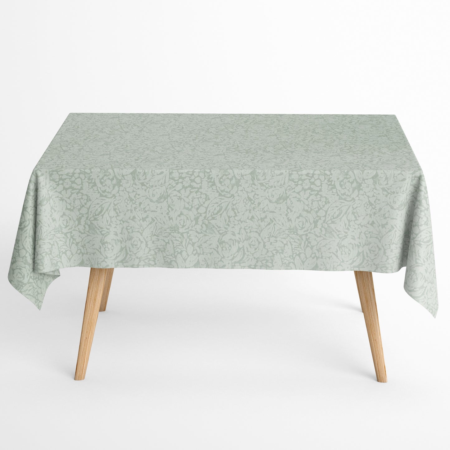 Beschichtete Baumwoll-Tischdecke in hellem Salbeigrün mit floralem Allover-Muster aus Blüten und Blättern auf hellem Hintergrund, über eine Tischecke drapiert. Die matte Oberfläche wirkt pflegeleicht, die Tischbeine aus hellem Holz sind sichtbar.