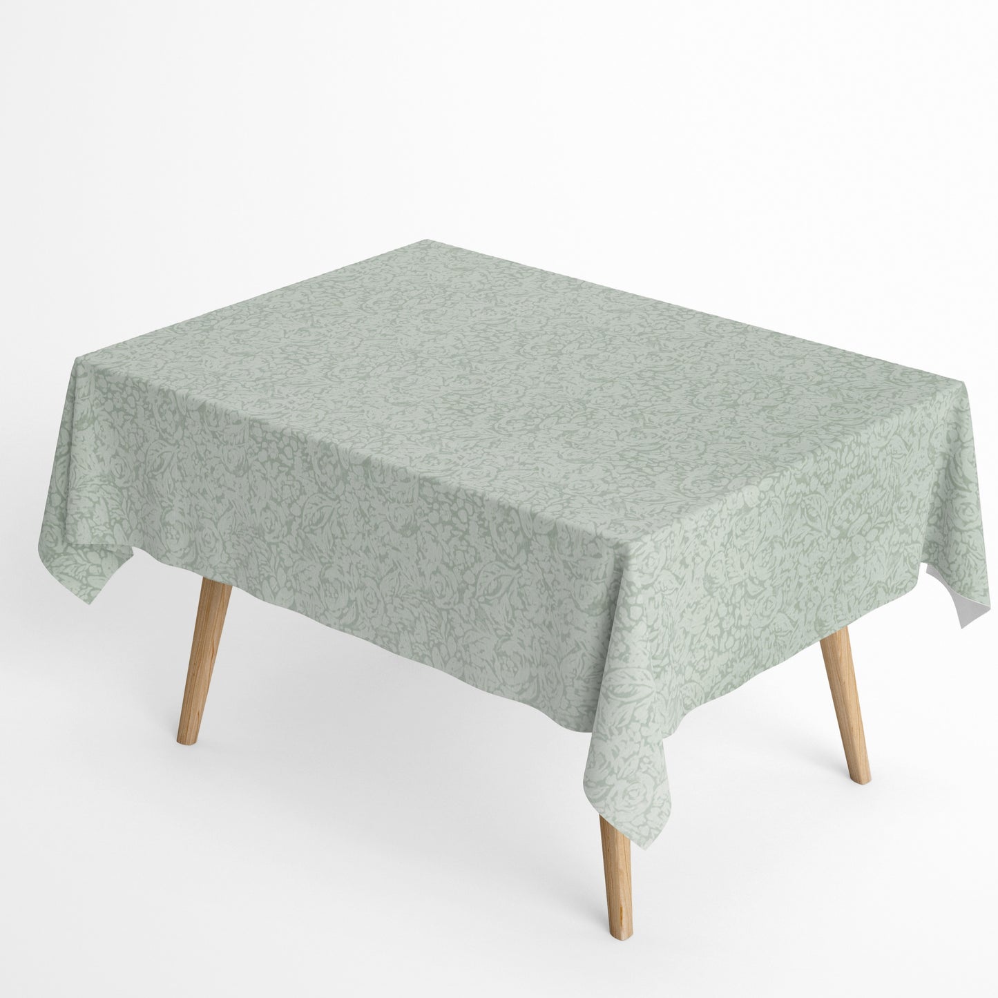 Beschichtete Baumwoll-Tischdecke in hellem Salbeigrün mit floralem Allover-Muster aus Blüten und Blättern auf hellem Hintergrund, über eine Tischecke drapiert. Die matte Oberfläche wirkt pflegeleicht, die Tischbeine aus hellem Holz sind sichtbar.