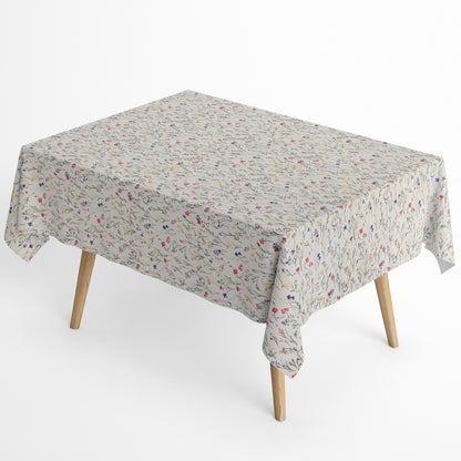Beschichtete Baumwoll-Tischdecke mit zartem Streublumenmuster auf hellem Hintergrund. Filigrane Wildblumen und Gräser in Rot, Blau, Gelb und Rosa verteilen sich gleichmäßig über die Oberfläche. Über eine Tischecke drapiert, mit sichtbaren hellen Holzbeinen.