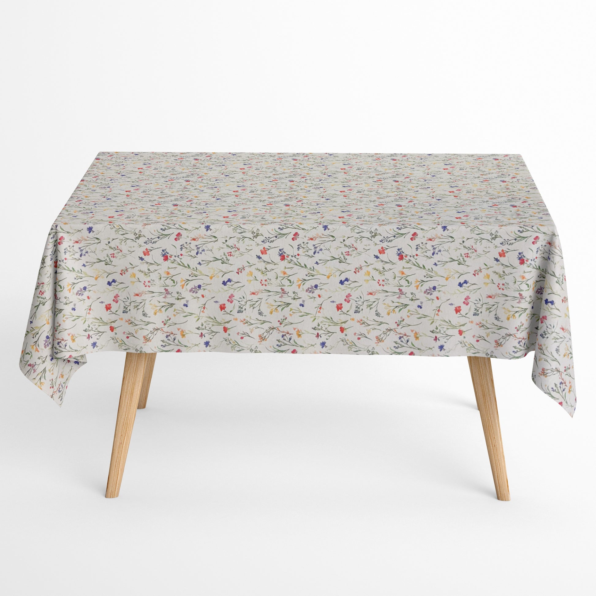 Beschichtete Baumwoll-Tischdecke mit zartem Streublumenmuster auf hellem Hintergrund. Filigrane Wildblumen und Gräser in Rot, Blau, Gelb und Rosa verteilen sich gleichmäßig über die Oberfläche. Über eine Tischecke drapiert, mit sichtbaren hellen Holzbeinen.