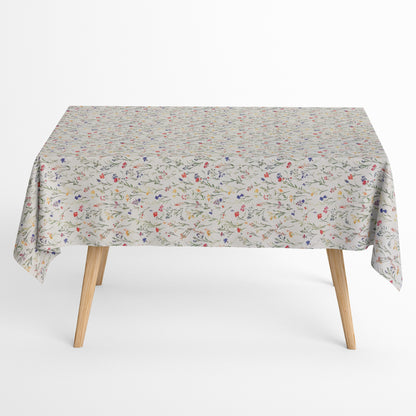 Beschichtete Baumwoll-Tischdecke mit zartem Streublumenmuster auf hellem Hintergrund. Filigrane Wildblumen und Gräser in Rot, Blau, Gelb und Rosa verteilen sich gleichmäßig über die Oberfläche. Über eine Tischecke drapiert, mit sichtbaren hellen Holzbeinen.