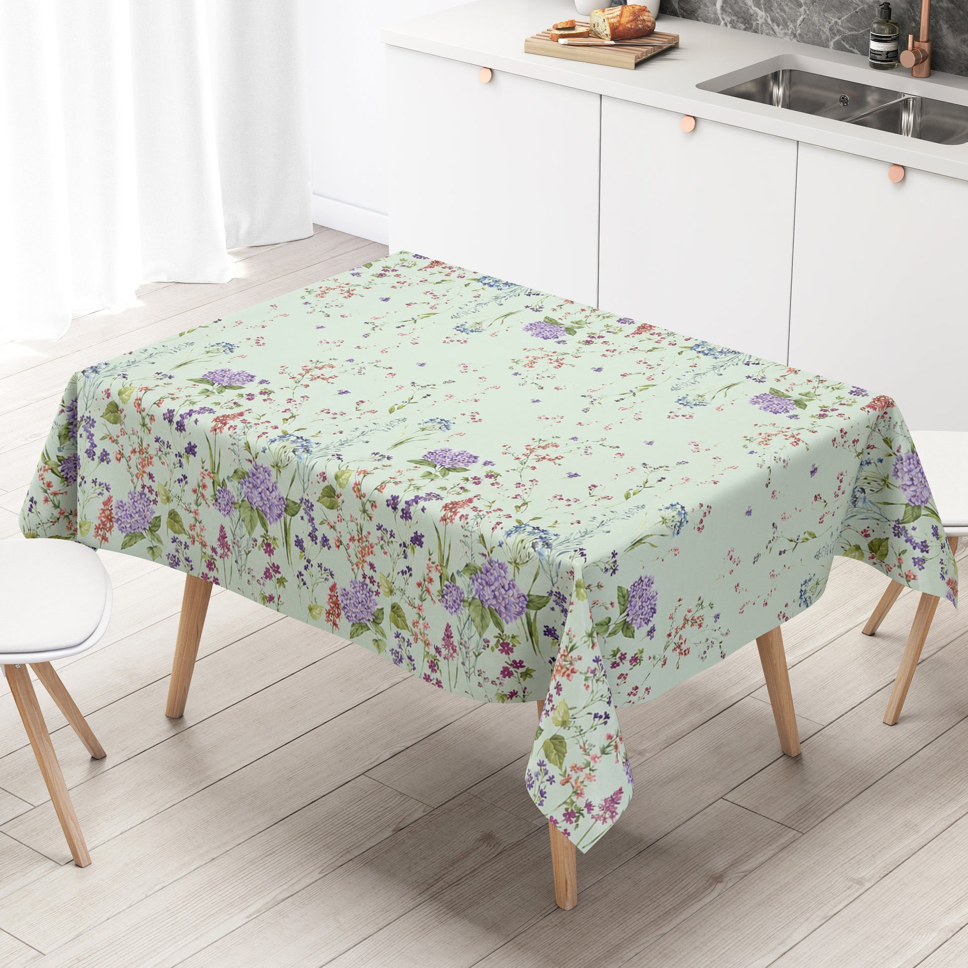 Wachstuchtischdecke mit romantischem Blumenmuster auf mintgrünem Hintergrund. Zarte Wildblumen, filigrane Zweige und Blütenranken in Rosa, Blau, Lila und Rot verteilen sich locker über die Fläche. Große violette Hortensien setzen dekorative Akzente entlang der oberen und unteren Bereiche. Das detailreiche, aquarellartige Dessin wirkt leicht, natürlich und verspielt und verleiht dem Tisch eine frische, florale Ausstrahlung.