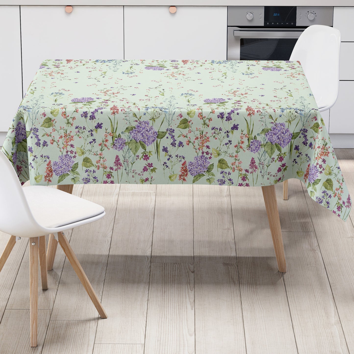 Wachstuchtischdecke mit romantischem Blumenmuster auf mintgrünem Hintergrund. Zarte Wildblumen, filigrane Zweige und Blütenranken in Rosa, Blau, Lila und Rot verteilen sich locker über die Fläche. Große violette Hortensien setzen dekorative Akzente entlang der oberen und unteren Bereiche. Das detailreiche, aquarellartige Dessin wirkt leicht, natürlich und verspielt und verleiht dem Tisch eine frische, florale Ausstrahlung.