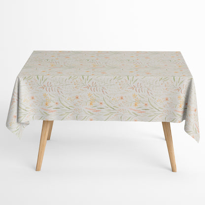 Beschichtete Baumwoll-Tischdecke mit filigranem Naturmuster auf hellem Hintergrund. Zarte Gräser, Blätter und kleine Blüten in Apricot-, Gelb- und Grüntönen verteilen sich gleichmäßig über die Oberfläche. Über eine Tischecke drapiert, mit sichtbaren hellen Holzbeinen.