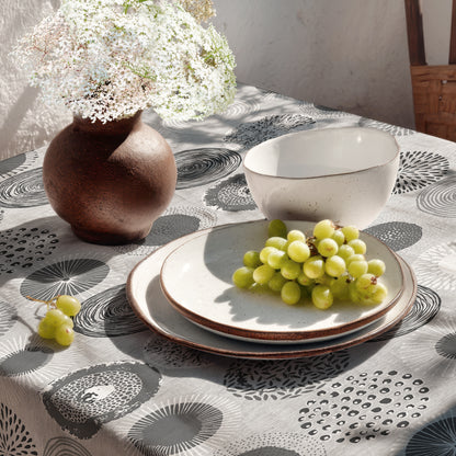 Jacquard-Tischdecke mit grafischem Kreismotiv aus Spiralen, Punkten, Strahlen und floralen Formen in Grau, Weiß und Anthrazit.