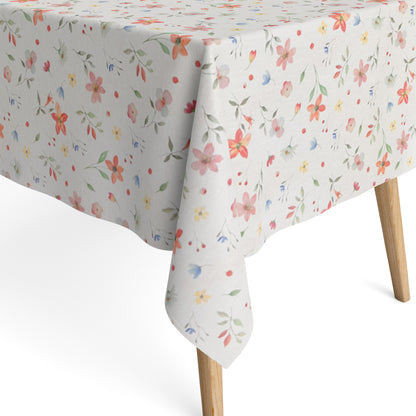 Beschichtete Baumwoll-Tischdecke mit zartem Streublumenmuster auf hellem Hintergrund. Kleine Blüten, feine Zweige und dekorative Punkte in Rosa, Korall, Gelb, Blau und Grün verteilen sich gleichmäßig über die Oberfläche. Über eine Tischecke drapiert, mit sichtbaren hellen Holzbeinen.
