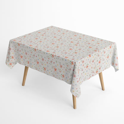 Beschichtete Baumwoll-Tischdecke mit zartem Streublumenmuster auf hellem Hintergrund. Kleine Blüten, feine Zweige und dekorative Punkte in Rosa, Korall, Gelb, Blau und Grün verteilen sich gleichmäßig über die Oberfläche. Über eine Tischecke drapiert, mit sichtbaren hellen Holzbeinen.