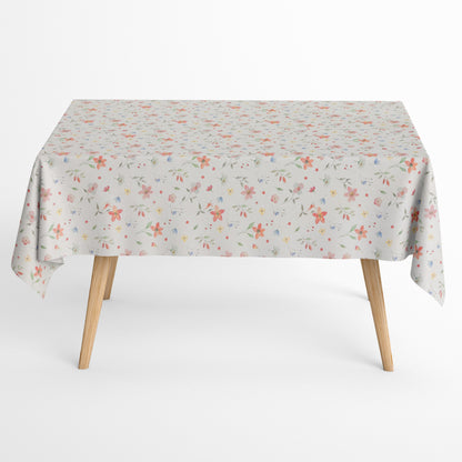 Beschichtete Baumwoll-Tischdecke mit zartem Streublumenmuster auf hellem Hintergrund. Kleine Blüten, feine Zweige und dekorative Punkte in Rosa, Korall, Gelb, Blau und Grün verteilen sich gleichmäßig über die Oberfläche. Über eine Tischecke drapiert, mit sichtbaren hellen Holzbeinen.